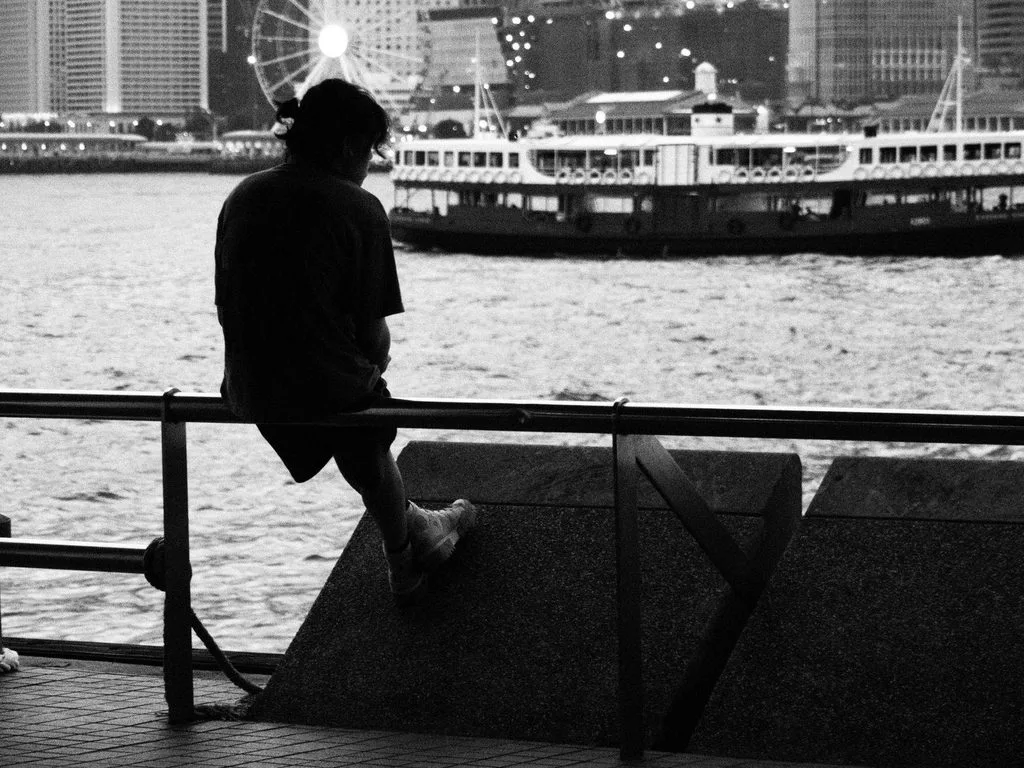 ilhouette of a person sitting by Hong Kong harbour as a ferry passes the skyline   