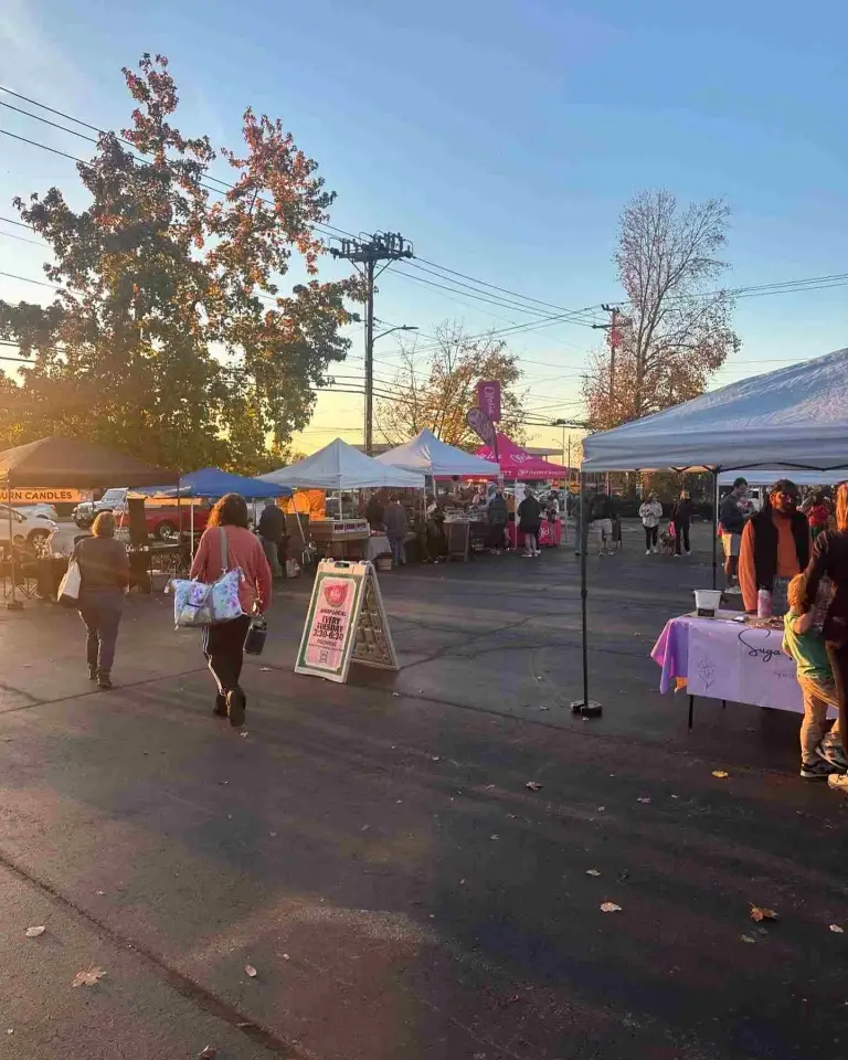 Vendors and customers mill around white tents at the East Nashville Farmers Market at sunset