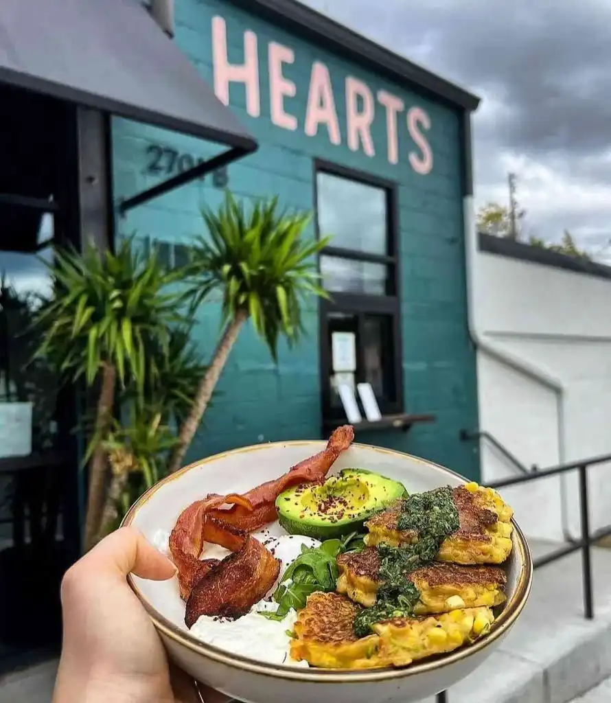 A hand holds a bowl of corn fritters, bacon, and avocado outside the teal Hearts Nashville exterior.
