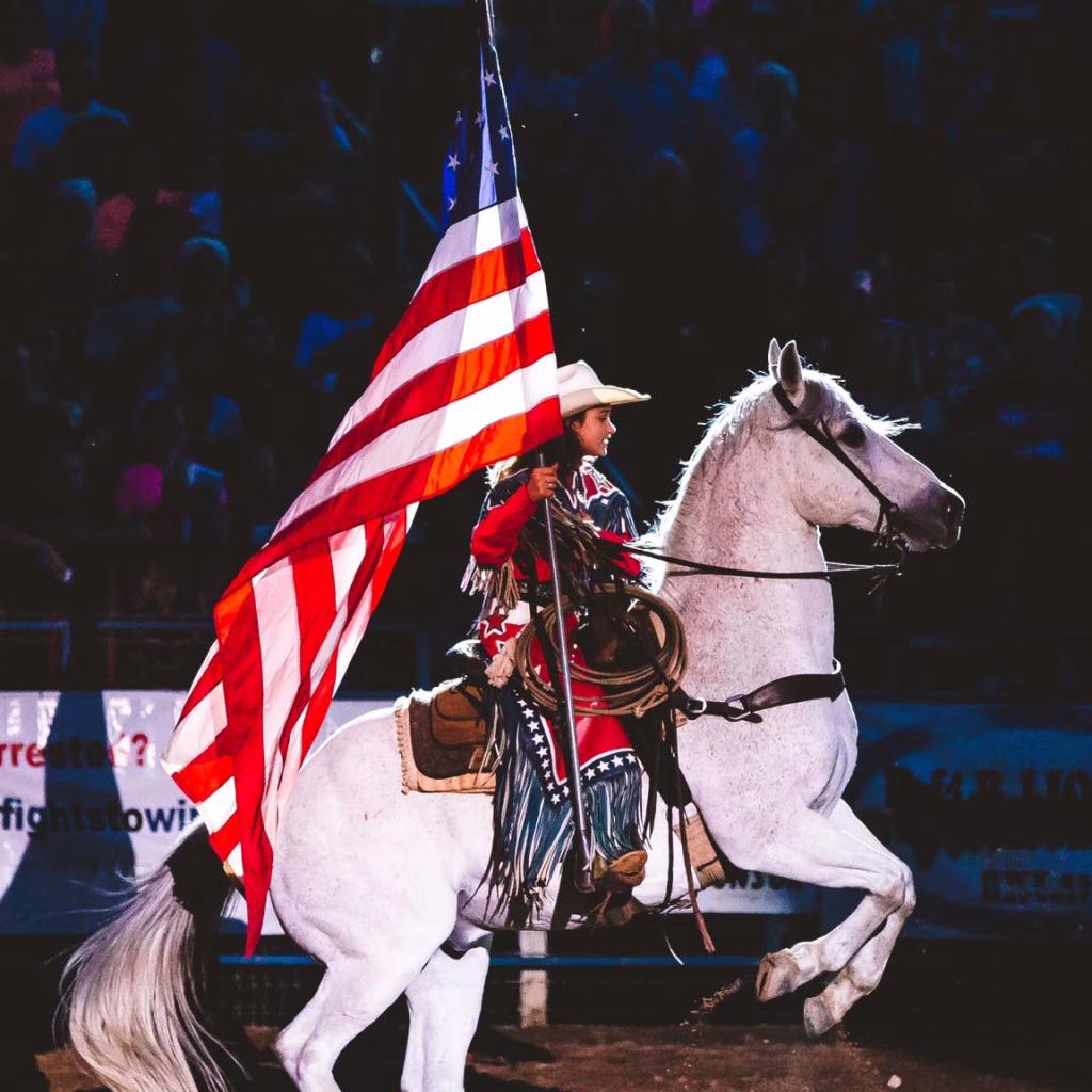 A cowgirl proudly rides her white horse carrying the American flag during a vibrant Nashville in July rodeo.