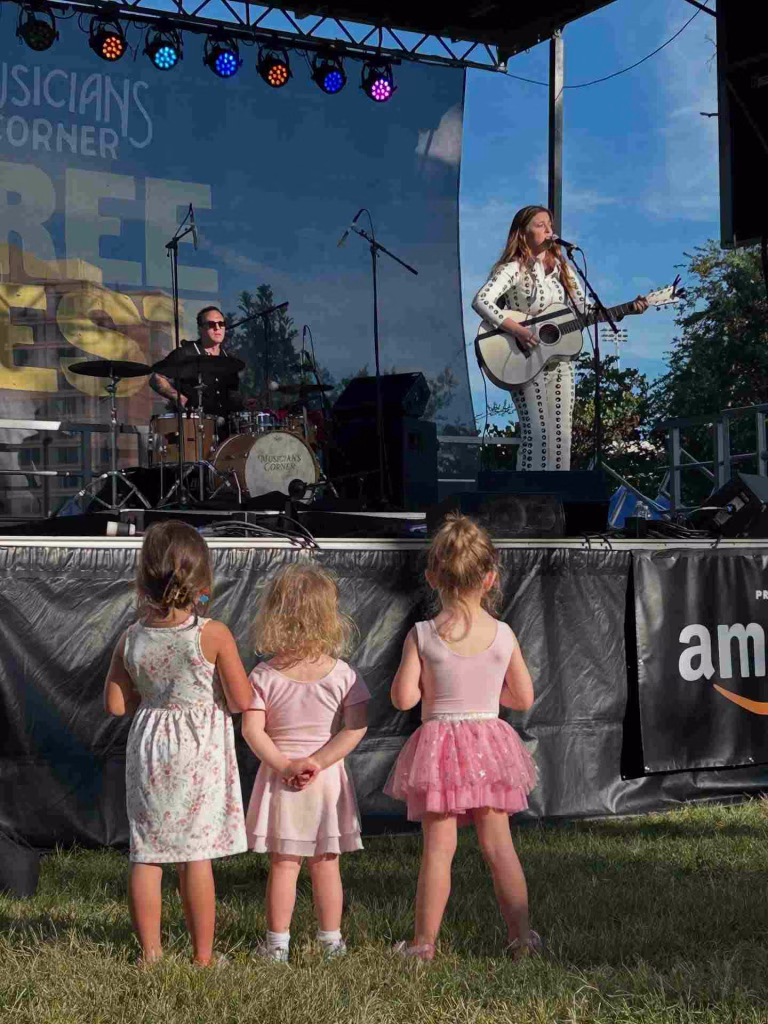 Three little girls in pink dresses watch a live outdoor concert in Nashville in September.