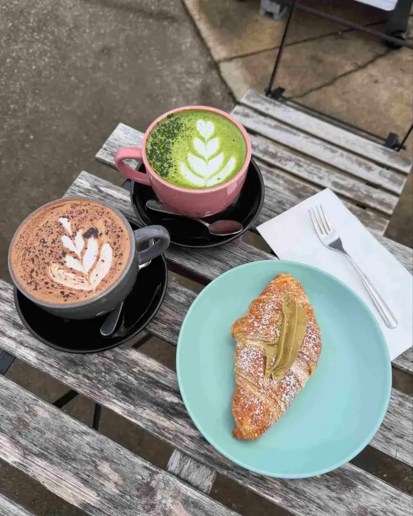 Two unique lattes and a croissant with green spread sit on a wooden table at All People Coffee Nashville