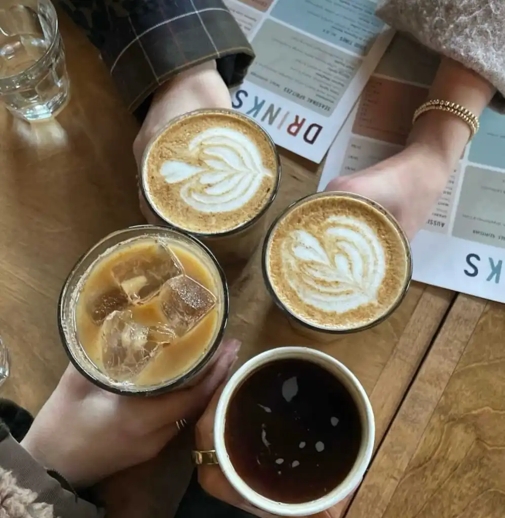 Four hands hold various coffee drinks (lattes, iced coffee, black) on a wooden table at Hearts Nashville.
