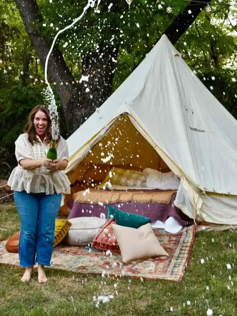 East Nashville Summer Bucket List Guide A woman smiling while shaking a champagne bottle, spraying foam outside a glamping tent for an East Nashville Summer Bucket List adventure.