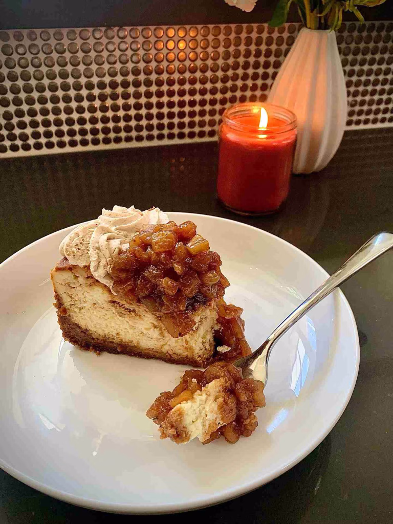 Slice of apple cheesecake on a white plate with a bite on a fork, a dessert on this East Nashville Guide.