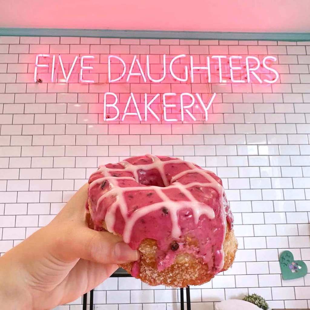 A hand holds a pink frosted donut in front of the Five Daughters Bakery neon sign in Nashville 12 South.