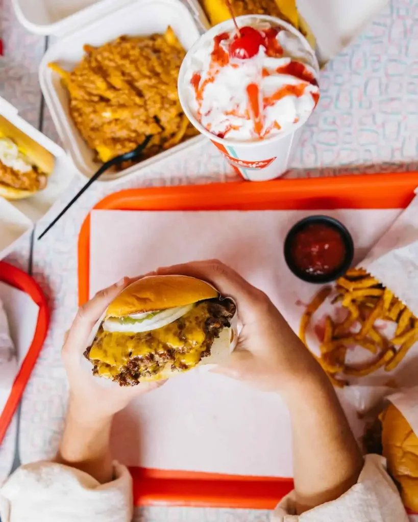 East Nashville Summer Bucket List Guide Hands holding a gooey cheeseburger, with fries and a milkshake, an essential East Nashville Summer Bucket List food item.